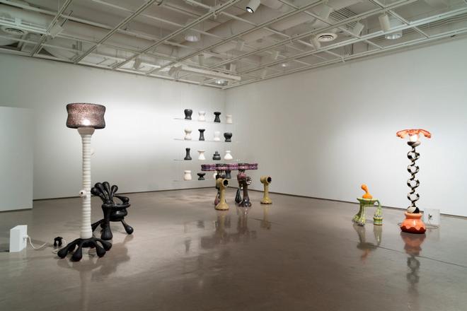 An installation view of the thesis exhibition. All made of wheel-thrown ceramics, featuring two lamps, a chair and ashtray, a small table, a bar and stools and a set of acrylic shelves with black and white forms sitting on them.