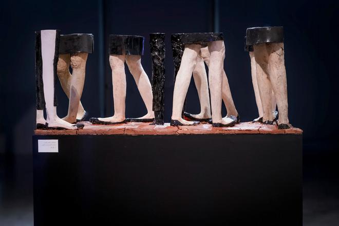 Sculpture of eight pairs of human legs from the thighs down, two wearing trousers with the other six wearing skirts, displayed on a dark platform with a layer of red clay, in a gallery setting.